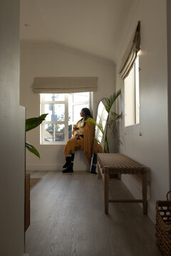 Indian woman sitting on multi-pane window ledge wearing mustard poncho holding ceramic mug in hall