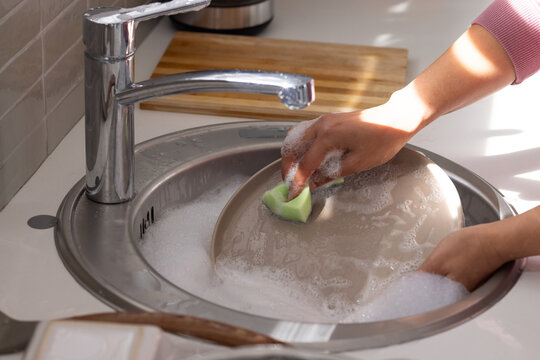 Large beige plate is being washed in stainless steel sink with green sponge and soap suds