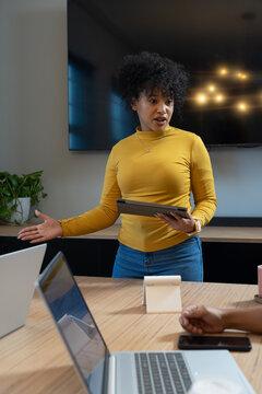 African American adult woman holding tablet and gesturing at office table with laptops, screen