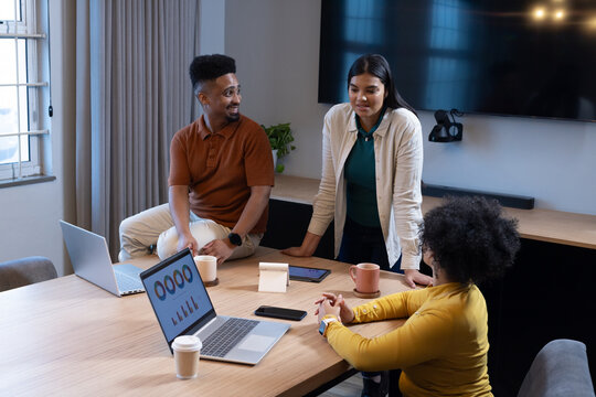 Diverse coworkers meeting at office wooden table in business attire, using laptops and coffee