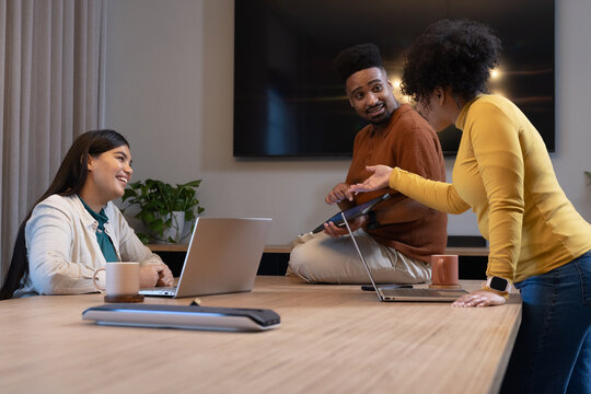 Diverse coworkers collaborating in conference room using laptops, tablet, coffee mugs