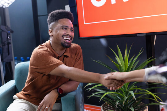 African American man smiling reaching shaking hands seated in armchair in studio near red monitor