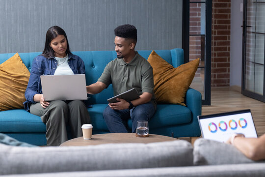 Diverse coworkers in denim and green polo collaborating on laptop and tablet in office lounge