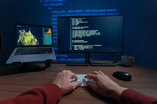 First person view of a programmer hands typing on a keyboard at night, two monitors displaying code and data science visualizations on dark desk, works on a machine learning or data science. POV