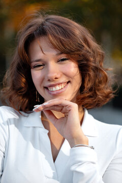Young woman smiling with hand beneath her chin in a white blouse during a sunny outdoor lifestyle portrait