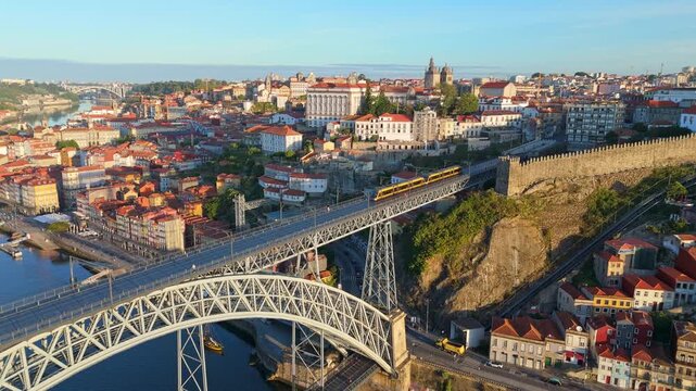 Drone view of Dom Luis I Bridge over Douro River in Porto, Portugal, with steel bridge arch, including a tram on the bridge and rabelo boats by the quay at sunrise. Orbit parallax shot