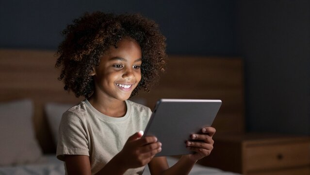 Young girl with curly hair happily engages with a tablet device on her bed at night, illuminated by soft screen glow.