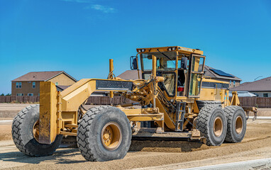 Fototapeta premium Heavy motor grader levels gravel for new roadway surrounded by residential construction. Land preparation.