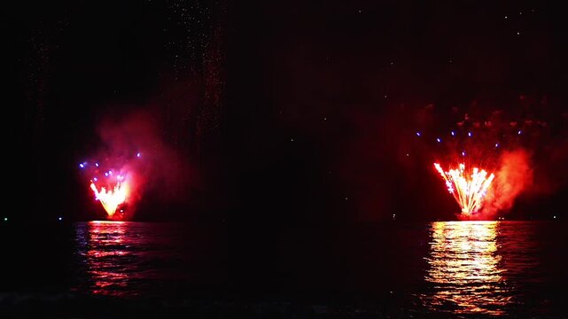 Distant view of bright red fireworks erupt from barges or shoreline launcher stations and reflect on the dark seawater surface at night.