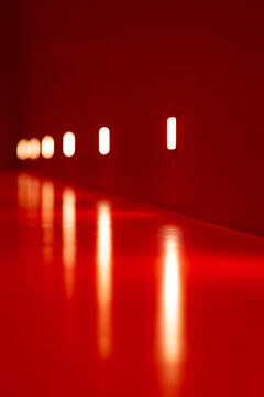 Minimalist surreal interior bathed in intense red light, with a rhythmic sequence of glowing white lamps and elongated reflections creating an abstract sense of depth and infinity.