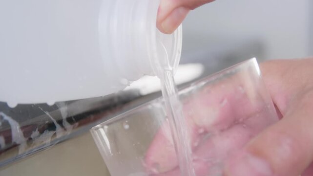 Close up of hands filling a glass of drinking water from a plastic dispenser on a kitchen countertop. Plastic tap is depressed and water is released.