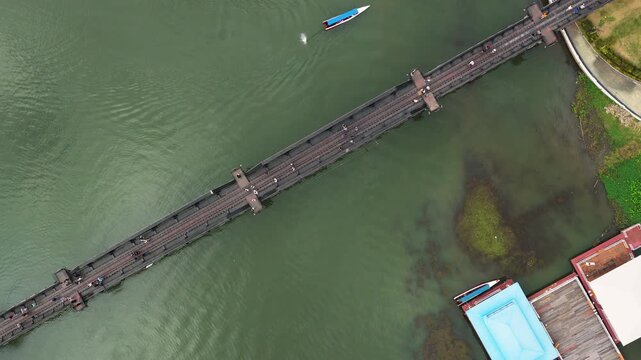 The steel bridge over the River Kwai cuts a diagonal across green water at Kanchanaburi, Thailand, visitors small on the deck and a long tail boat passing below. Aerial top down flyover