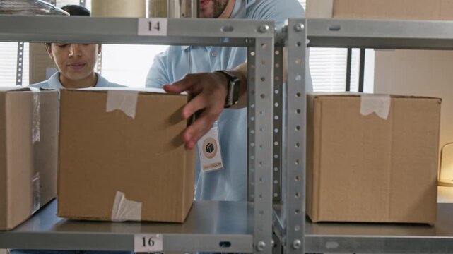 Caucasian young man placing cardboard boxes on metal shelf while female coworker checks inventory using digital tablet. Perfect for e-commerce logistics and supply chain marketing