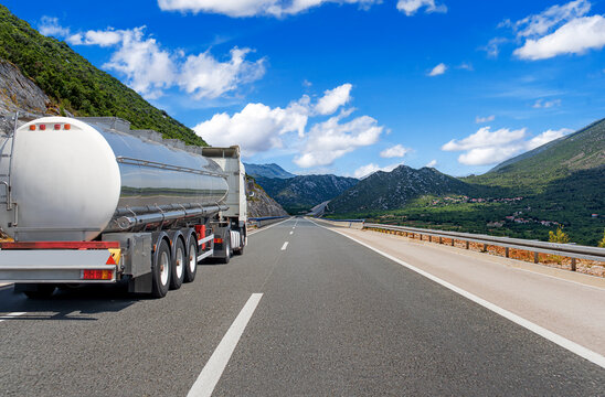 A silver tanker truck drives along an open highway through a scenic mountain landscape under a bright blue sky.