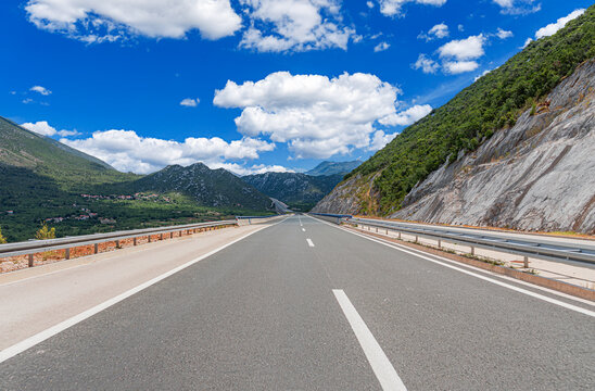 An empty highway stretches through a scenic mountain landscape under a bright blue sky with fluffy white clouds.