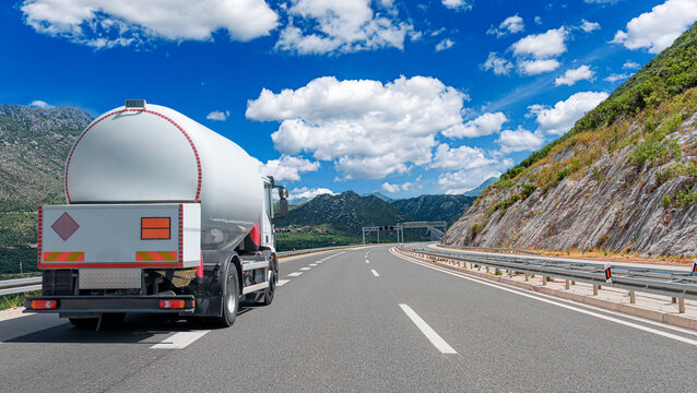 A white tanker truck drives along a mountain highway under a blue sky with fluffy clouds.