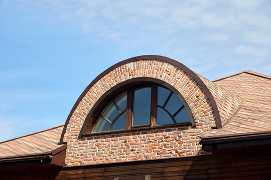 An arched dormer window featuring a semi-circular brick surround, multi-pane glass design, brown wooden framing, and textured roof shingles under a clear blue sky.