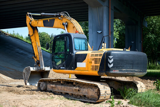Yellow tracked excavator parks on dirt ground under concrete bridge structure. Heavy construction machinery with bucket stands near embankment on industrial building site.