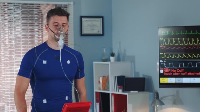 An adult male wearing a royal blue athletic shirt stands indoors with oxygen mask and electrodes connected, monitored by medical equipment in a clinical laboratory.