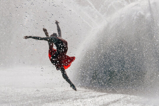 White dancer leaping through water droplets in a red dress outdoors