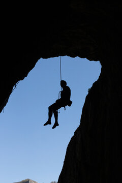 Silhouette of a rock climber rappelling down from a high cave opening against a clear blue sky
