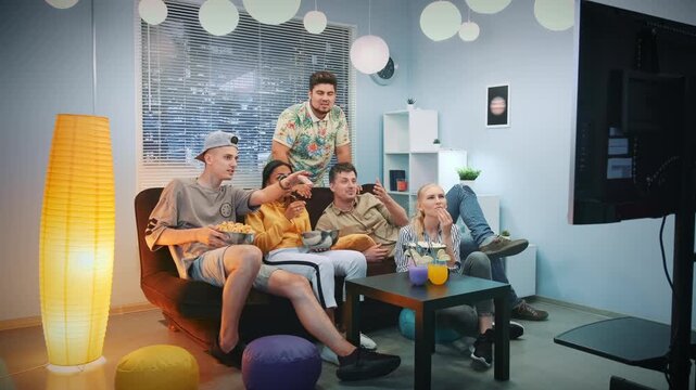 Six adults casually dressed are seated on a sofa watching a large TV and eating snacks in a bright, modern indoor living room during daytime.