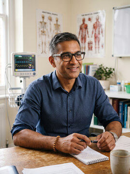 Friendly male doctor sitting at his desk in a modern consultation room, illustrating approachable primary healthcare and patient trust.