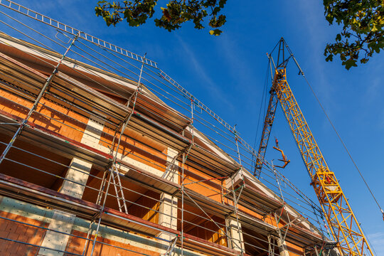 Low angle view of modern building under construction with metal scaffolding and yellow crane under clear blue sky, residential development and construction site concept