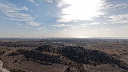 Fototapeta premium Aerial view of dry agricultural terrain in Cyprus with rocky hills, cultivated fields and distant coastal plains beneath a bright sky with scattered clouds