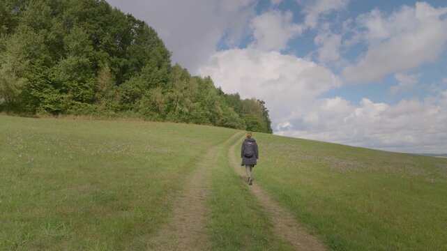 A woman with a backpack is walking uphill along a grassy slope, with an open landscape and clouds in the background. A trip into nature