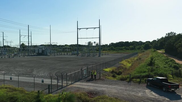 Two workers in hi vis stand at the gate of an electrical substation at Gentry, Arkansas, razor wire along the fence and steel gantries rising over the switchyard. Aerial view