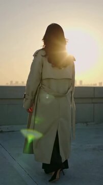 Vertical video: Walking woman in trench coat, heels pacing rooftop parapet as sun sinks, flaring