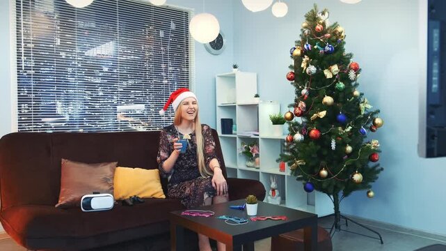 A smiling woman in a patterned dark dress and red Santa hat sits on a sofa holding a blue cup in a modern living room decorated with a Christmas tree and city skyscrapers visible outside.