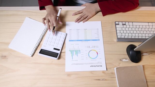 Female professional accountant conducting detailed financial analysis, meticulously reviewing budget reports with charts and calculator on wooden workplace surface.