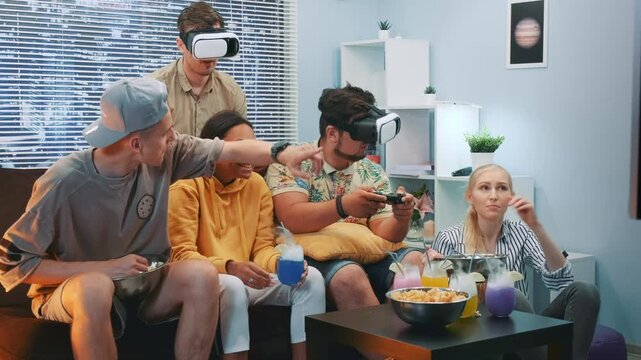 Six adults, three men and three women, play virtual reality games while sitting on a sofa in a bright, modern living room with snacks and drinks on the table.