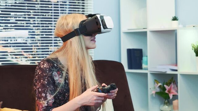 A blonde woman wearing a patterned top is smiling while using a black and white virtual reality headset and controller indoors on a dark brown sofa.