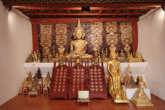 A golden seated Buddha statue centered before gilded floral stenciled wall panels on an elevated sanctuary altar inside Wat Phra That Chae Haeng Phra Aram Luang in Nan, Thailand.