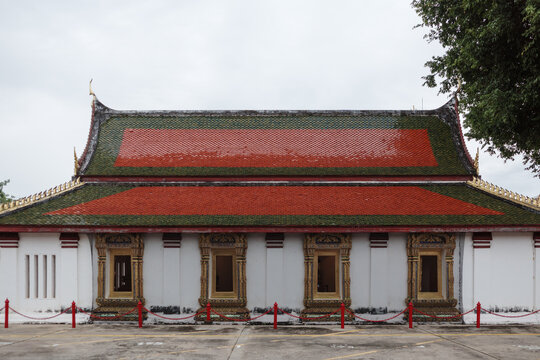 Symmetrical exterior eye-level shot of a white temple hall featuring ornate carved gilded window frames and a two-tiered green and orange glazed ceramic tiled gabled roof at Phuttha Chinnarat temple