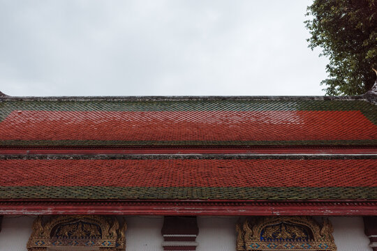 Low-angle outdoor eye-level shot of the multi-tiered red and green ceramic tiled roof and ornate golden window lintels of a traditional Buddhist temple building beneath an overcast sky.  