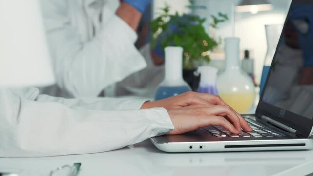 Scientist wearing white lab coat typing on a laptop in a chemistry laboratory with colorful flasks and a colleague nearby at a white desk during daytime.