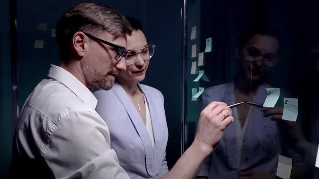 Business professionals brainstorming new ideas and strategy, collaborating on project development using sticky notes on a glass board in a dark modern office meeting room