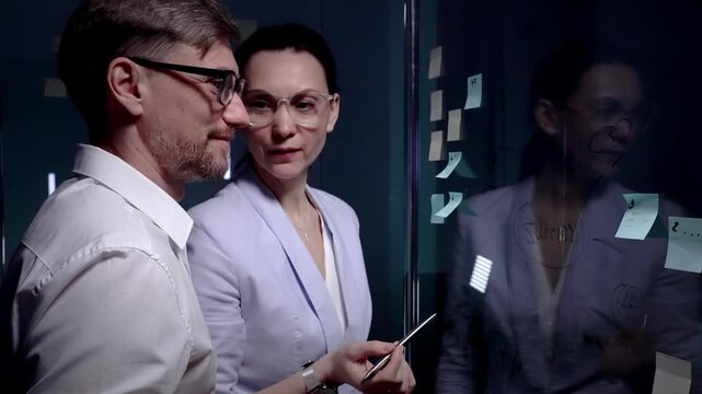Business professionals brainstorming new ideas and strategy, collaborating on project development using sticky notes on a glass board in a dark modern office meeting room