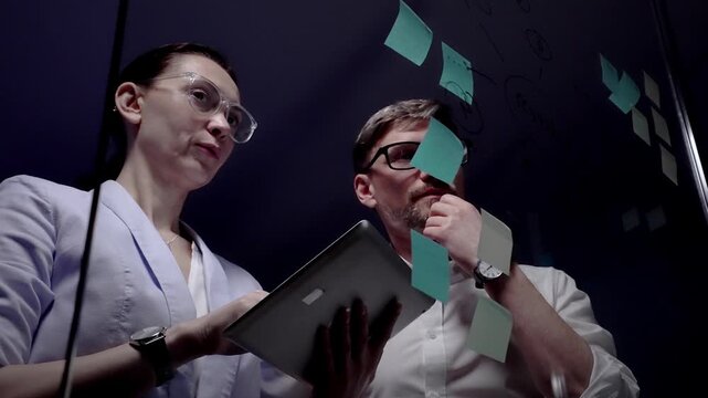 Business colleagues collaborating on new innovative project, drawing mind map with success word on clear glass writing board with sticky notes in modern office environment