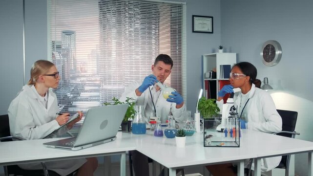 Three scientists in lab coats collaborate on a chemical experiment using a conical flask in a modern laboratory with laptops and scientific equipment.