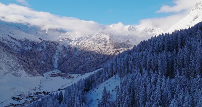 Aerial footage of snowy alpine peaks in Austria with a glacier rising through morning mist, snowfall. Blue sky, sunlight, and conifer forests in the foreground create a peaceful winter atmosphere.