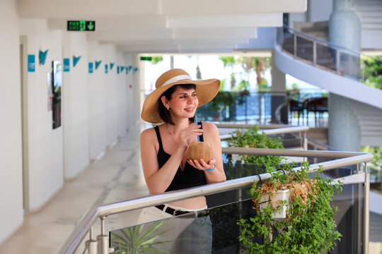 Woman in hat and black top standing hotel holding coconut with straw, tropical vacation drink, resort lifestyle, summer holiday concept.