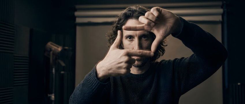 Professional film director, making framing gesture with hands looking at camera, curly hair male creator in dark sweater with equipment, cinematography studio background, for creative media marketing