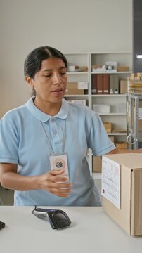 Vertical video of Hispanic young female worker scanning barcode on cardboard box and giving parcel to client. Clip portrays shipping logistics, mail fulfillment, and customer service