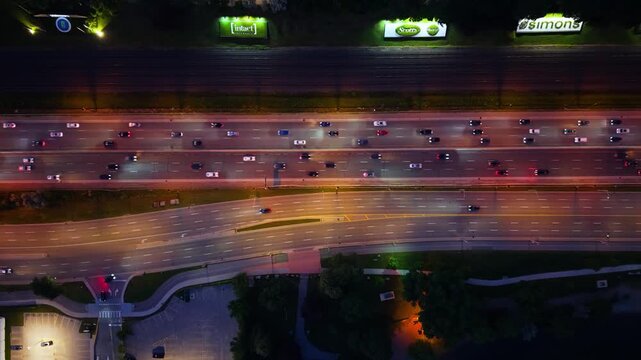 Overhead night highway interchange with dense traffic, curving ramp, glowing streetlights, reflective asphalt, roadside billboards, parking area, and urban transport atmosphere
