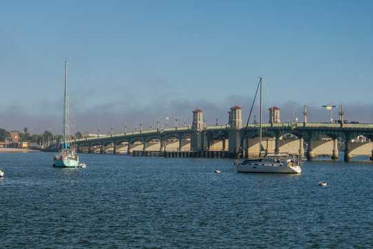 Two sailboats rest on blue water near the Bridge of Lions in Saint Augustine, Florida. The historic arched drawbridge stretches across the harbor beneath a clear sky.